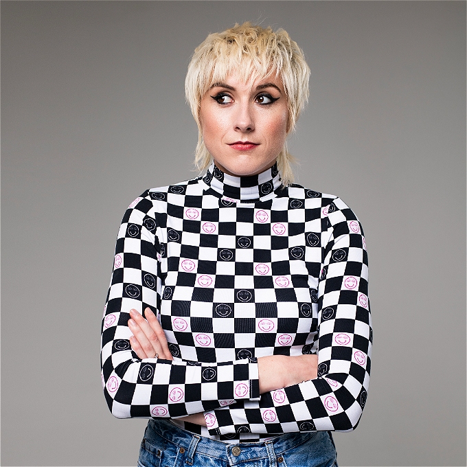 Comedian Maisie Adam is posing with her arms crossed, giving a side-eyes look, wearing a turtle neck jumper with a chess board pattern and white and pink smiley faces.