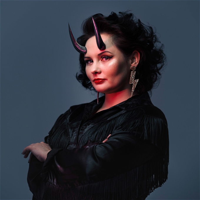 Rachel Fairburn is stood in front of a grey background. She is wearing a black jacket with tassels, lightning bolt earrings and has purple horns coming out of her forehead. The lighting is dramatic and moody.