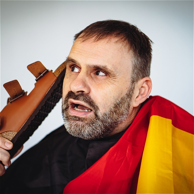 A promotional image for Jürgen Strack's comedy show featuring him listening to his Birkenstock sandal as though it's a phone. The background includes a blue sky and the text 'ACHTUNG!! The Germans Are Coming! Das Second Coming.