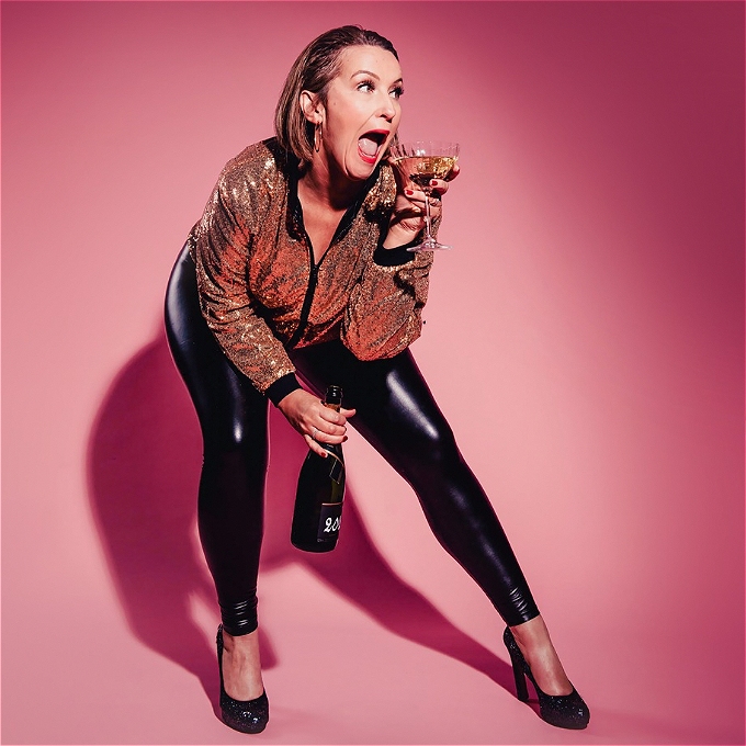 Teresa, a woman with short brown hair wearing a gold sequined top and black leather trousers, standing in front of a pink background. She is holding a glass of champagne in one hand, and a bottle in the other.