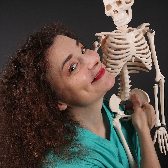 Female comedian wearing surgical scrubs laughing holding a small plastic skeleton