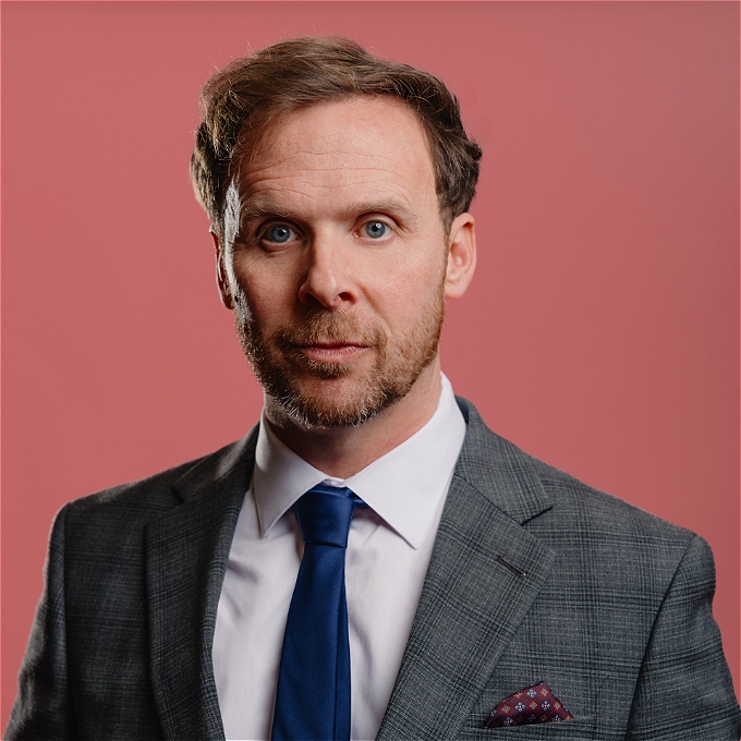 Colin Hoult wears a neutral expression a suit jacket, with pocket square, white shirt and blue silk tie against a pink background