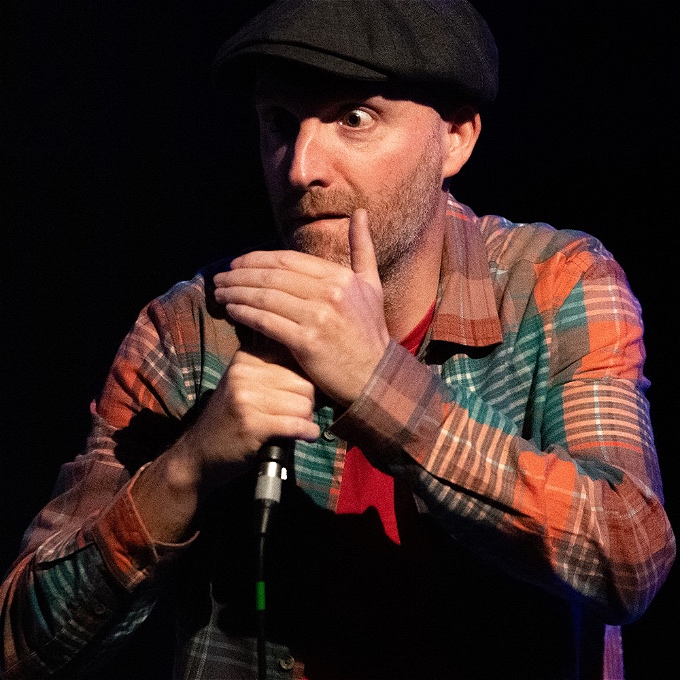 Male comic storyteller, mic in hand, wearing a flat cap