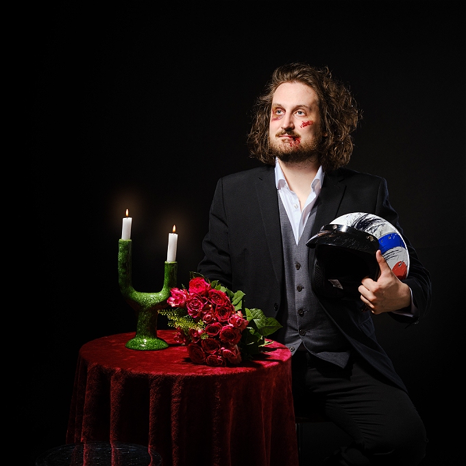 A man with long brown hair sits at a table set up for a date, he is bruised and holding flowers and a battered motorcycle helmet