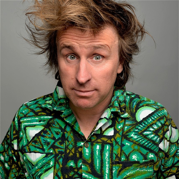 Milton Jones (comedian) with brown hair and wearing a patterned green shirt looking directly at the viewer.